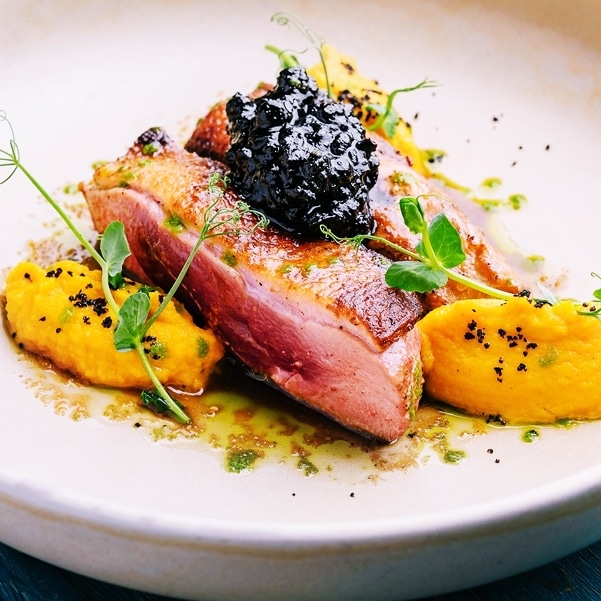 Rosa gebratene Entenbrust, serviert mit Cassis-Sauce und cremigem Süßkartoffel-Sellerie-Püree, dekoriert mit frischen Kräutern.